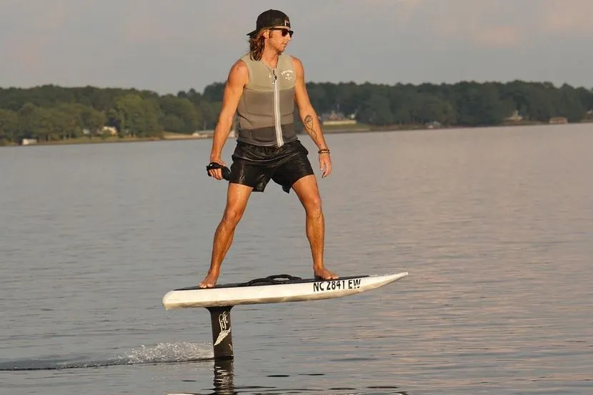 A rider standing on an eFoil crossing flat water on a lake, holding a wireless throttle, with vessel registration numbers printed on the nose of the board.